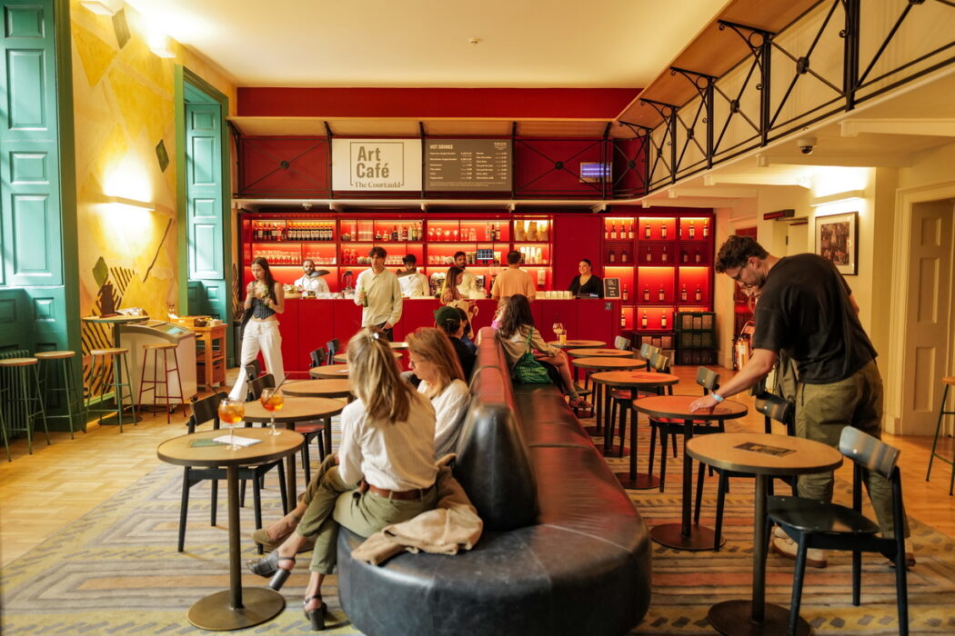 Interior of a modern Art Cafe with a vibrant red backdrop. The cafe features a long red bar with shelves of bottles, round wooden tables with black chairs, and a large curved gray seating area in the foreground. Customers are seated throughout the space, and staff members are working behind the bar. Green-framed windows and warm yellow walls add color to the contemporary cafe design. A mezzanine level with black metal railings is visible in the background.