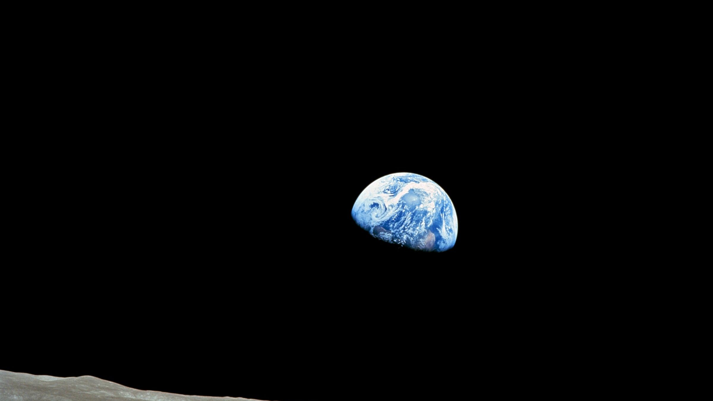 A photograph of Earth from lunar orbit, showing the small blue planet suspended in the vast darkness of space. The foreground features the Moon’s barren, beige surface, contrasting with the deep black sky and blue Earth.